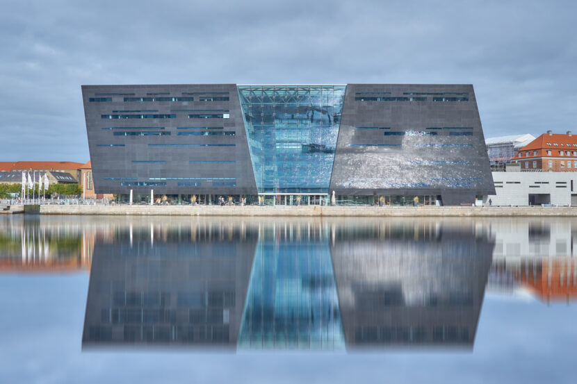 Die Königliche Bibliothek „Black Diamond“ in Kopenhagen mit Spiegelung im Wasser unter bewölktem Himmel