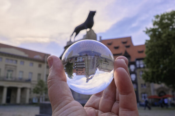 Das Foto zeigt den Burgplatz, den zentralen Platz, von Braunschweig in Niedersachsen. In der Mitte des Platzes steht auf einem Sockel eine Bronzeplastik des Braunschweiger Löwen. Das Bild ist durch einen Lensball, eine Glaskugel, hindurch fotografiert. Daher steht der Löwe etwas verkleinert in der Glaskugel auf dem Kopf, ist aber schwarf. Der eigentliche Löwe ist im Hintergrund des Bildes unschwarf zu erkennen.
