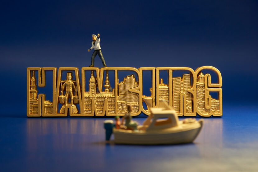Dies ist ein Bild mit Miniaturfiguren. Auf dem Foto ist ein Kühlschrank-Magnet mit dem Schriftzug "Hamburg" zu sehen. Auf dem Magneten steht ein winkender Matrose. Vor dem Schriftzug fährt ein Motorboot vorbei.