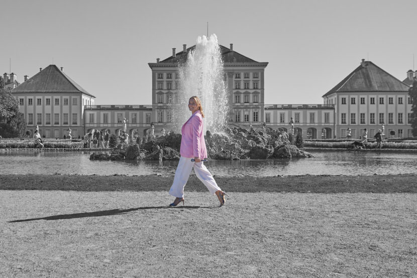 Auf dem Bild ist die Münchner Sängerin Sandra Weiß zu sehen. Im Hintergrund ist das Schloss Nymphenburg. Mittig im Hintergrund ist ein Springbrunnen. Die Sängerin geht von rechts nach links durch das Bild. Das Foto zeigt die Sängerin auf Höhe des Springbrunnens. Sie schaut zur Kamera und hält in der der Kamera zugewandten Hand ein Mikrofon. Das Bild an sich ist schwarz-weiß, nur Sandra Weiß selbst ist farbig. Markant ist ihr rosafarbener Blazer.
