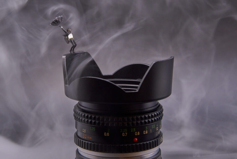 Dies ist ein Bild mit Miniaturfiguren. Zu sehen ist ein Mann mit einem Regenschirm. Die Fläche des Regenschirms ist wie von heftigen Regen nach oben gebogen. Der Mann steht auf dem Rand Objektivring eines Fotoobjektivs. Um ihn herum sind Nebelschaden zu sehen. Der Mann wirkt, als würde er da am Rand des Objektivs gleich das Gleichgewicht verliehren.