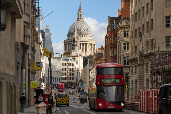 Ein roter Londoner Doppeldeckerbus fährt durch die Straßen. Im Hintergrund sieht man St. Paul’s Cathedral. Historische Fassaden und moderne Gebäude prägen die typische Londoner Stadtansicht bei Tageslicht.