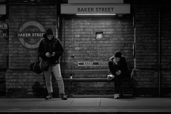 Schwarz-weiß Foto, das an der U-Bahn-Station Baker Street in London aufgenommen wurde. Das Bild zeigt zwei wartende Männer vor einer auf der Central Line typischen Backsteinwand der U-Bahn-Station, die auf ihre Handys schauen, während sie auf die U-Bahn warten.