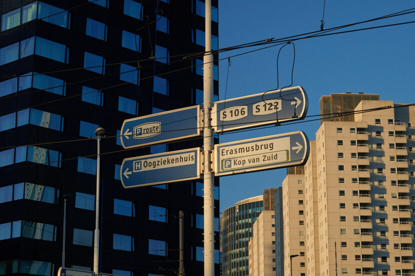 Wegweiser in Rotterdam vor moderner Hochhauskulisse mit Hinweisen zur Erasmusbrücke und Kop van Zuid