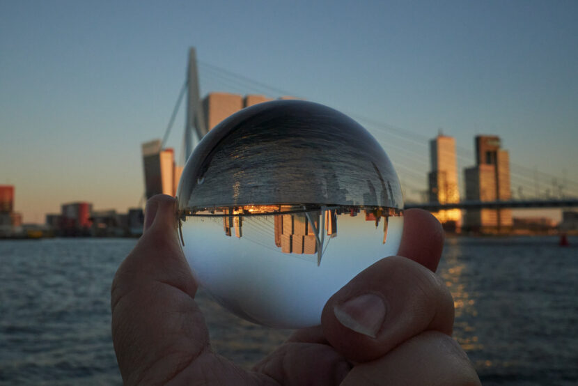 Glaskugel mit spiegelverkehrter Skyline von Rotterdam und Erasmusbrücke im Abendlicht am Wasserufer