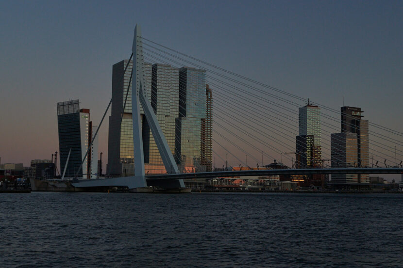 Die Erasmusbrücke in Rotterdam spannt sich bei Abendlicht über die Maas. Moderne Hochhäuser spiegeln die urbane Skyline wider, während das ruhige Wasser die Szene ergänzt.