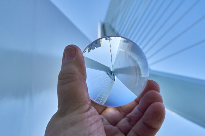 Eine Hand hält eine Glaskugel vor die Erasmusbrücke in Rotterdam. Die filigranen Seile und Linien der modernen Brücke spiegeln sich klar in der Kugel vor hellem Himmel wider.