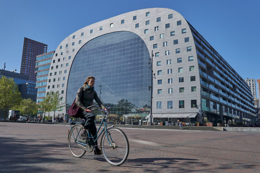 Eine Radfahrerin fährt vor der modernen Markthal in Rotterdam vorbei. Das markante Gebäude mit großer Glasfront und bogenförmiger Architektur prägt den urbanen Platz unter blauem Himmel.