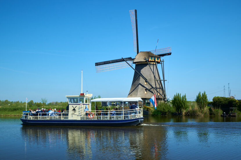 Ein Ausflugsboot fährt auf einem Kanal in Kinderdijk nahe Rotterdam an einer historischen Windmühle vorbei. Die UNESCO-Welterbestätte zeigt typisch niederländische Landschaft unter blauem Himmel.